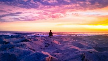 A gorgeous pink purple, and orange sunset at a white sand beach on the ocean with a silhouette of an observer sitting in the sand.