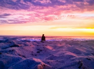 A gorgeous pink purple, and orange sunset at a white sand beach on the ocean with a silhouette of an observer sitting in the sand.