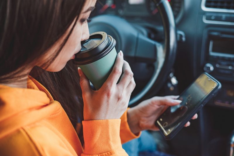 A woman drinks her coffee and checks her phone in her car before getting on the highway