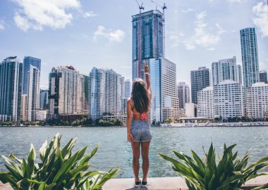 Woman in jean shorts and a swimsuit top faces the skyline of Miami and gives the peace sign across the water.