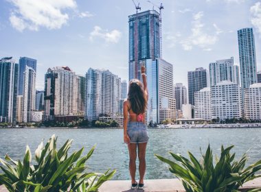 Woman in jean shorts and a swimsuit top faces the skyline of Miami and gives the peace sign across the water.