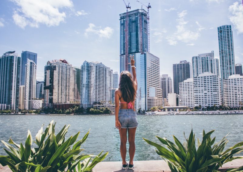 Woman in jean shorts and a swimsuit top faces the skyline of Miami and gives the peace sign across the water.