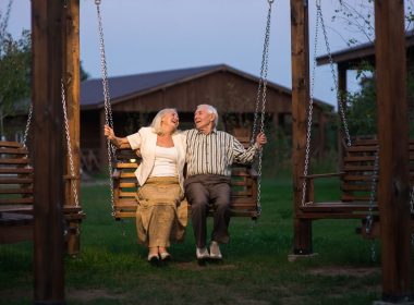 An old couple smiles at each other while sitting on a wooden swing at dusk.