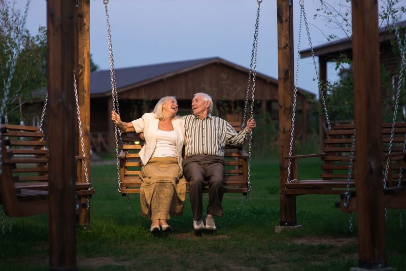 An old couple smiles at each other while sitting on a wooden swing at dusk.