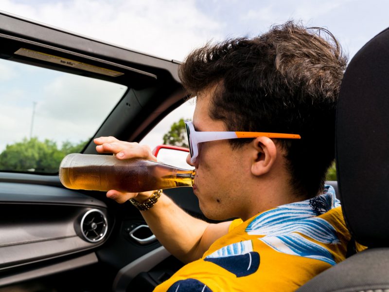 A man takes a swig of beer in the pasenger seat of a convertible as it drives down the road.