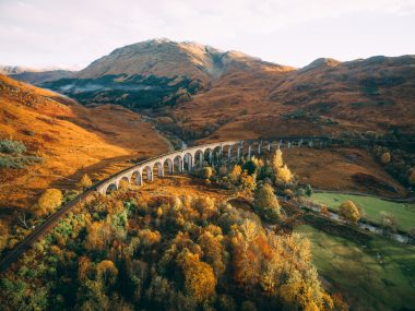 Glenfinnan Viaduct, Fort William, Scotland. Aerial view of the river and bright orange and green foliage in the warm glow of an autumn sunset.
