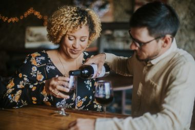 A man pours wine for a woman while they sit together in a winery.