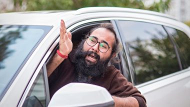 A man with long hair, a beard, and glasses leans out of his car window to yell at something in front of him.