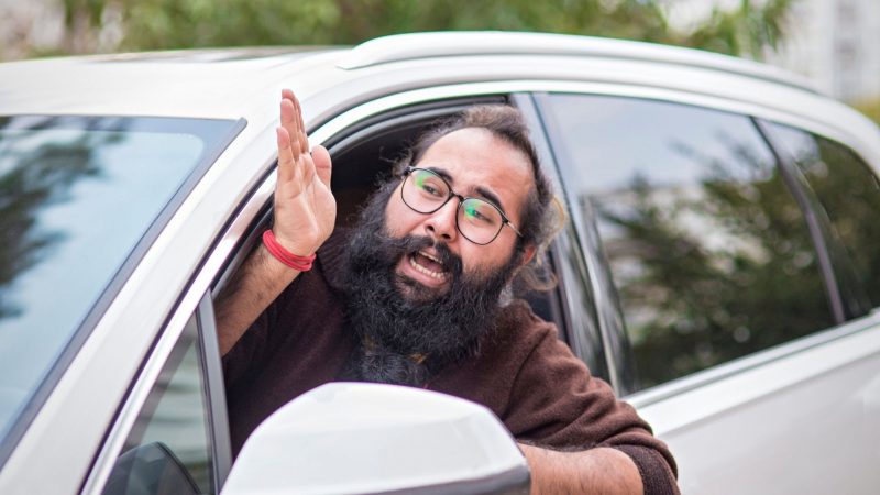 A man with long hair, a beard, and glasses leans out of his car window to yell at something in front of him.
