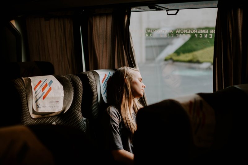 A woman leans her head towards the window to sleep while she travels on a bus.