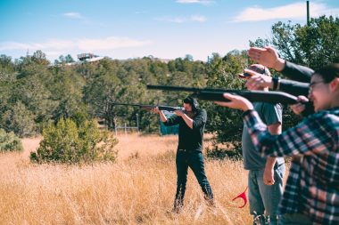 A group of people hold up their shotguns and aim at clay pigeons flying through a field.