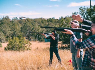 A group of people hold up their shotguns and aim at clay pigeons flying through a field.