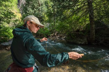 A woman in a hat, sunglasses, and jacket readies to cast as she fly fishes a creek in the woods.