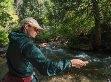 A woman in a hat, sunglasses, and jacket readies to cast as she fly fishes a creek in the woods.