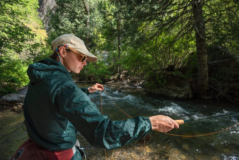 A woman in a hat, sunglasses, and jacket readies to cast as she fly fishes a creek in the woods.