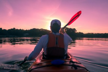 A kayaker's back as he paddles across a glass river towards the sunset that has turned the sky and water pink
