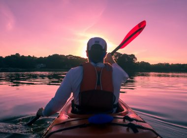 A kayaker's back as he paddles across a glass river towards the sunset that has turned the sky and water pink