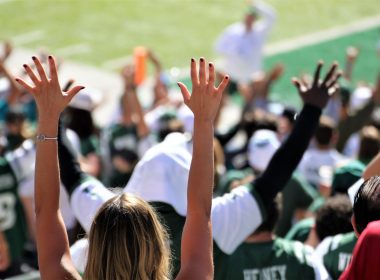 Cheering fans with their hands up in the stands of a football game.