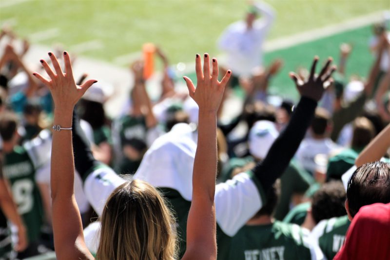 Cheering fans with their hands up in the stands of a football game.