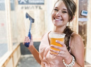 A smiling woman holds a beer and an axe at an Axe Throwing facility.