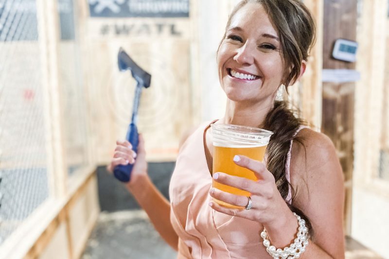 A smiling woman holds a beer and an axe at an Axe Throwing facility.