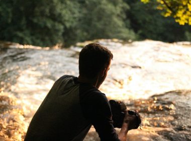 A man in the shadow of trees crouched near a river flowing into a small waterfall.