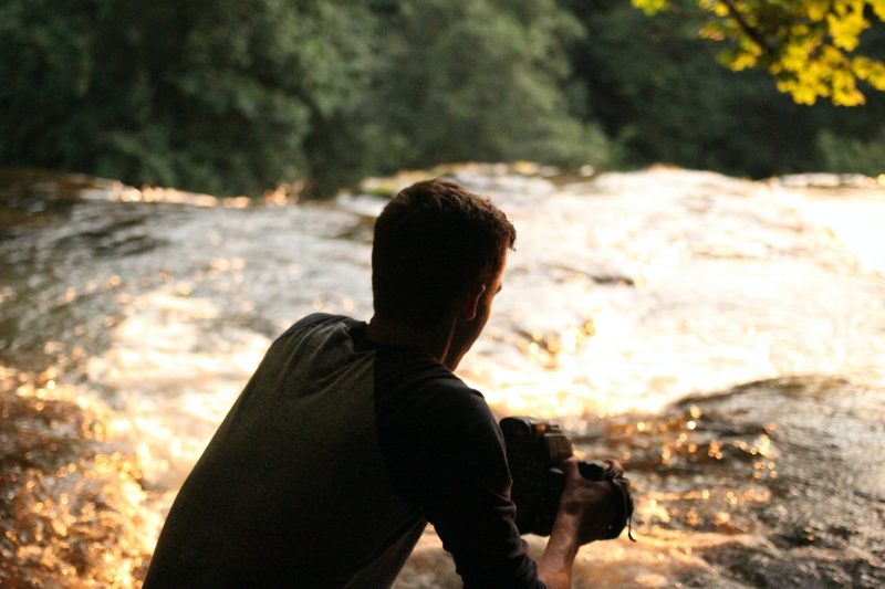 A man in the shadow of trees crouched near a river flowing into a small waterfall.