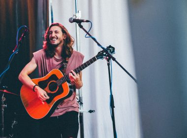 A musician performs on stage with his guitar and grins out to the audience