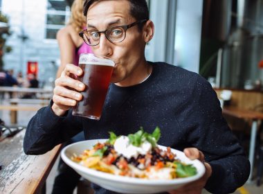 A man drinks a craft beer at a brewery and holds up a plate of nachos.