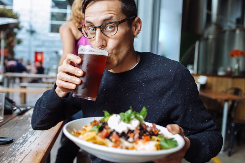 A man drinks a craft beer at a brewery and holds up a plate of nachos.