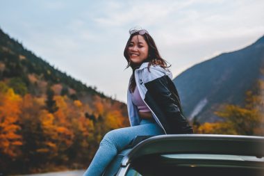 A young woman sits on top of her car as she stops during a fall road trip