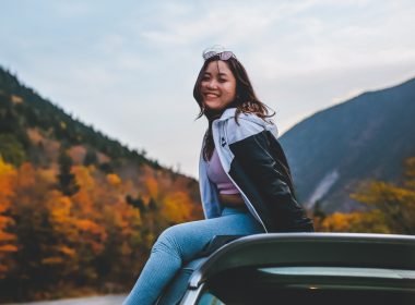 A young woman sits on top of her car as she stops during a fall road trip