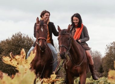 Two people horseback riding in the fall on brown horses.