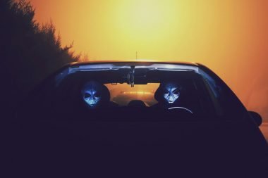 Two aliens with their faces illuminated by the light of the dashboard drive a car at night.