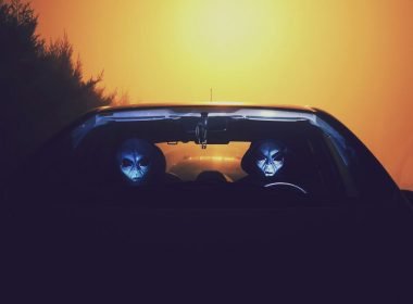 Two aliens with their faces illuminated by the light of the dashboard drive a car at night.