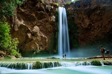 A tall waterfall cascading over red rocks in Arizona, creating a bright blue pool of water below where visitors dip their toes.
