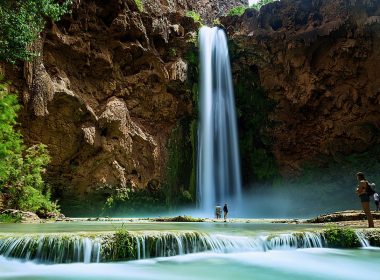 A tall waterfall cascading over red rocks in Arizona, creating a bright blue pool of water below where visitors dip their toes.