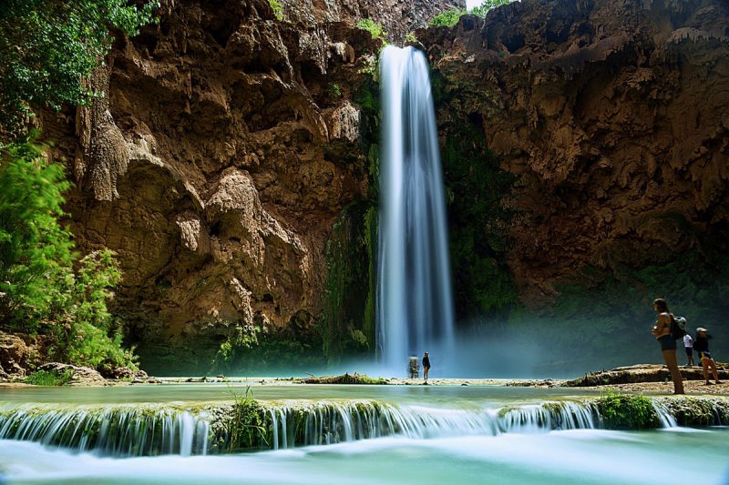 A tall waterfall cascading over red rocks in Arizona, creating a bright blue pool of water below where visitors dip their toes.
