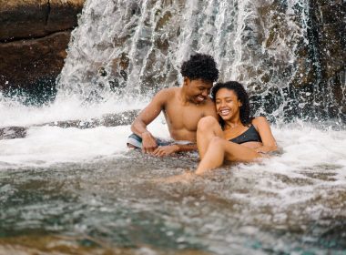 A couple sits in the creek, laughing and swimming at the bottom of a small waterfall.