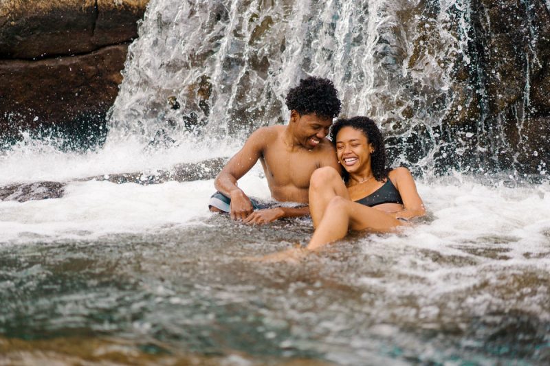 A couple sits in the creek, laughing and swimming at the bottom of a small waterfall.