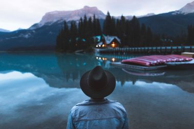 An explorer looks across a lake at a row up canoes and a warmly lit cabin.