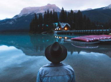 An explorer looks across a lake at a row up canoes and a warmly lit cabin.
