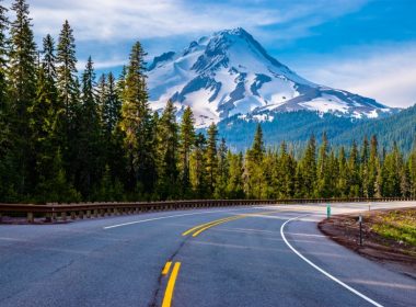 Driving down a paced road toward Mount Hood as it towers over the landscape, covered in snow.