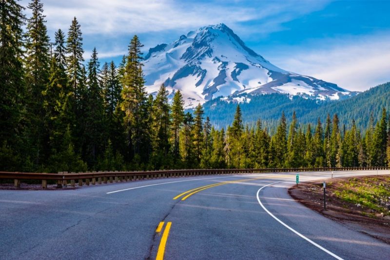 Driving down a paced road toward Mount Hood as it towers over the landscape, covered in snow.