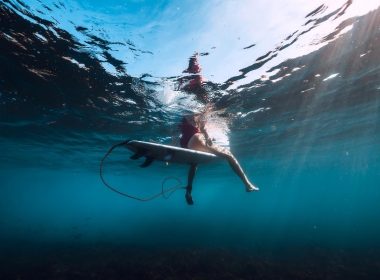 From below the water's surface, a sufer's legs dangle precariously of her surfboard in the depths of the ocean.