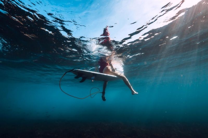 From below the water's surface, a sufer's legs dangle precariously of her surfboard in the depths of the ocean.