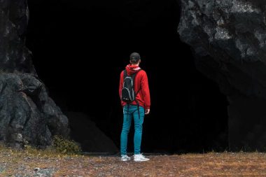 A young man with a backpack stands in font of a dark mine entrance.