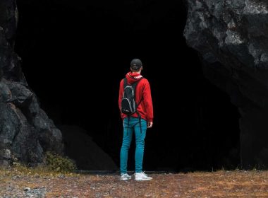 A young man with a backpack stands in font of a dark mine entrance.