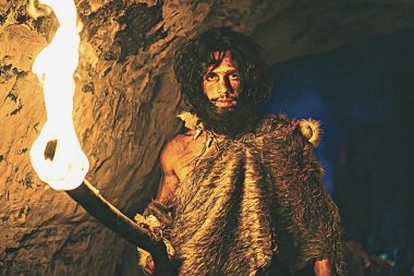 A depiction of an ancient cave man with dark red hair and a flaming torch in a dark cave.