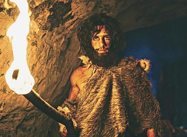 A depiction of an ancient cave man with dark red hair and a flaming torch in a dark cave.